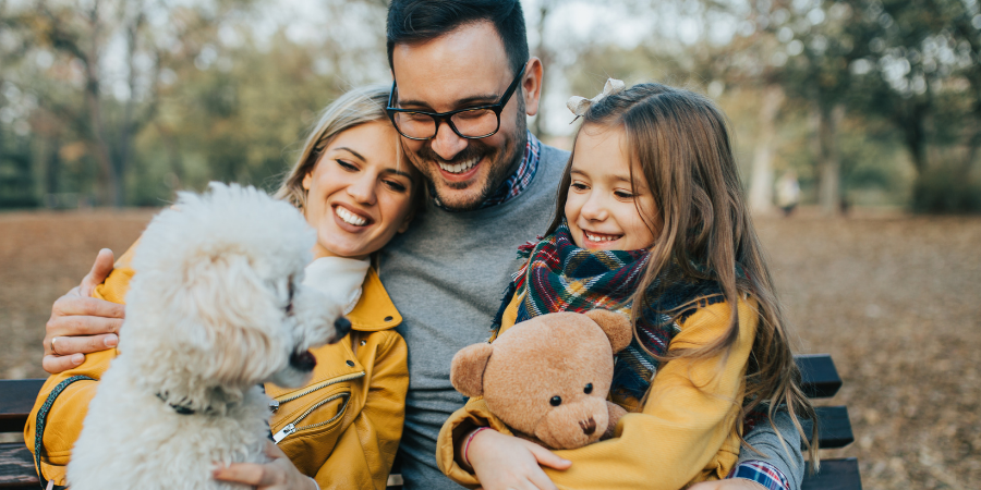 A smiling family sits on a park bench with a white fluffy dog and a teddy bear during autumn.