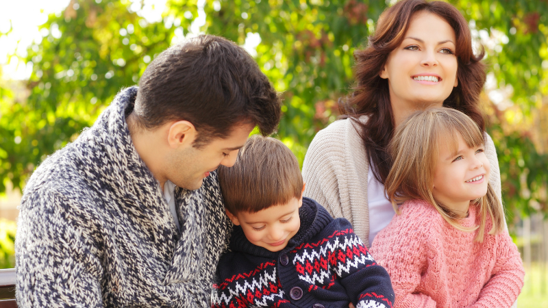 A family of four sits on a park bench smiling, with a father and son on the left and a mother with a daughter on the right outdoors.