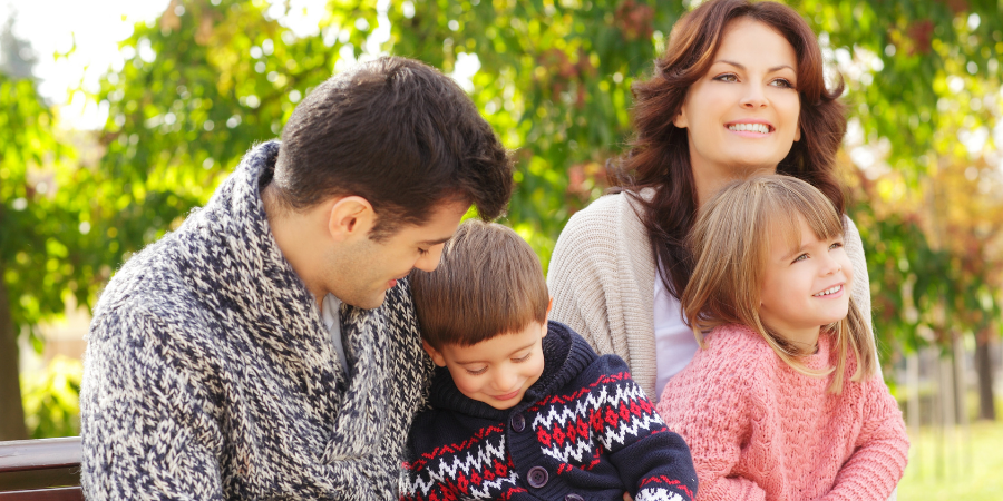 A family of four sits on a park bench smiling, with a father and son on the left and a mother with a daughter on the right outdoors.