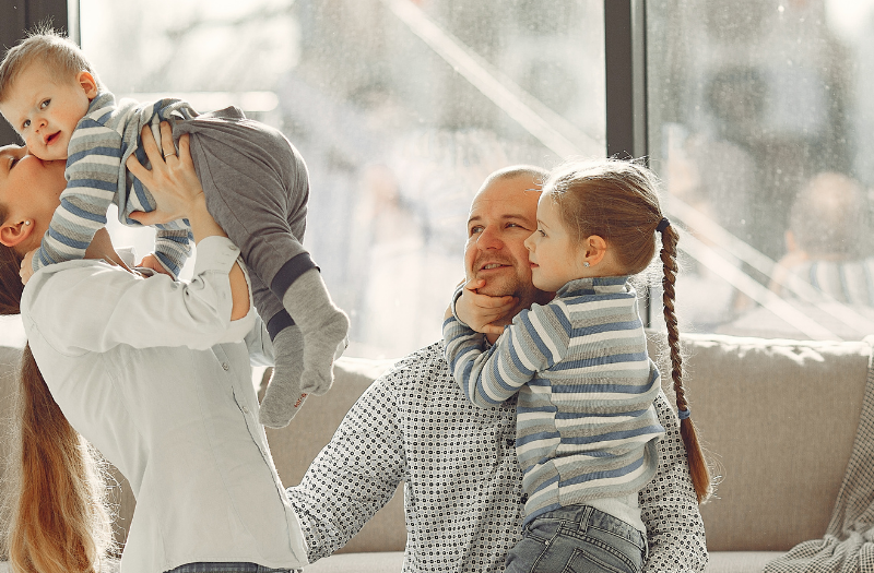 Parents playfully lift baby while seated on a couch in a sunlit living room, two kids smiling nearby.