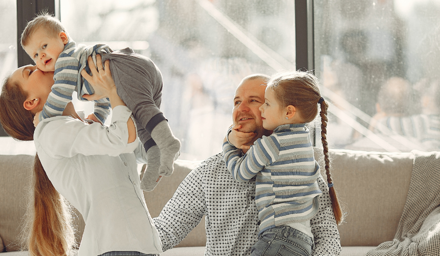 Parents playfully lift baby while seated on a couch in a sunlit living room, two kids smiling nearby.