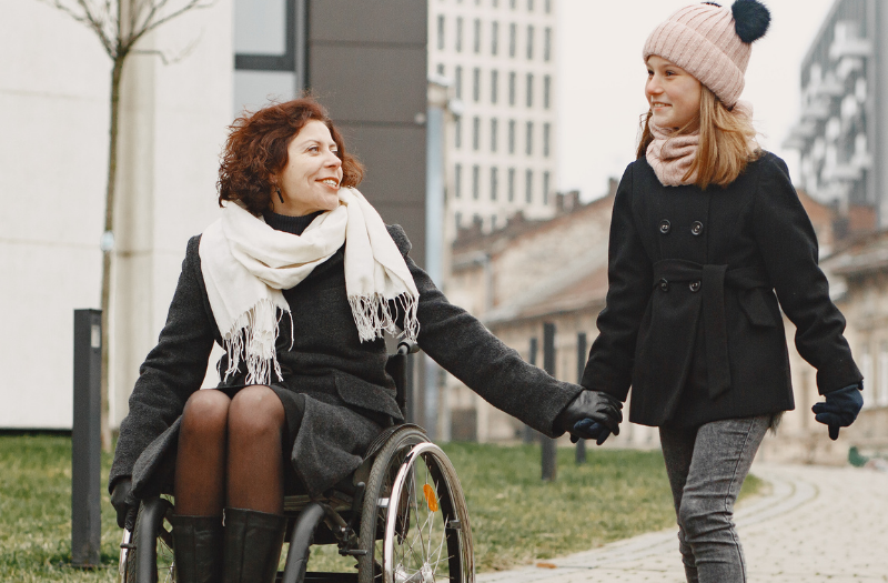 Woman in a wheelchair and a girl walking hand in hand along a city path in winter.