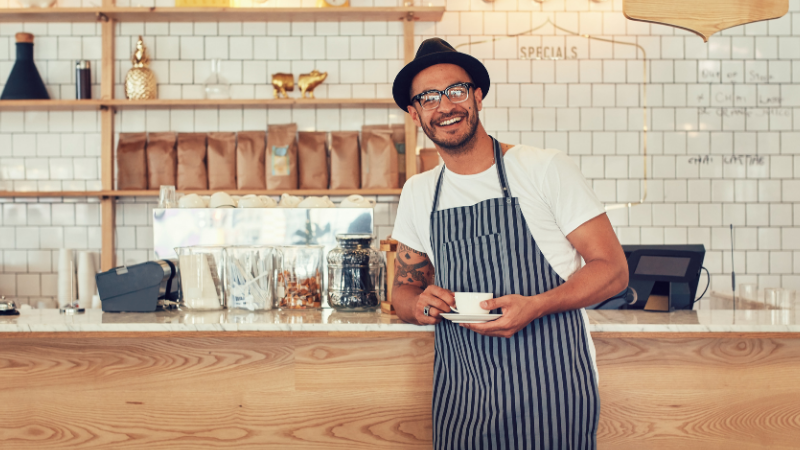 Barista in a striped apron standing behind a wooden counter, holding a cup and smiling in a coffee shop.