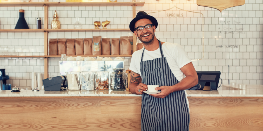 Barista in a striped apron standing behind a wooden counter, holding a cup and smiling in a coffee shop.