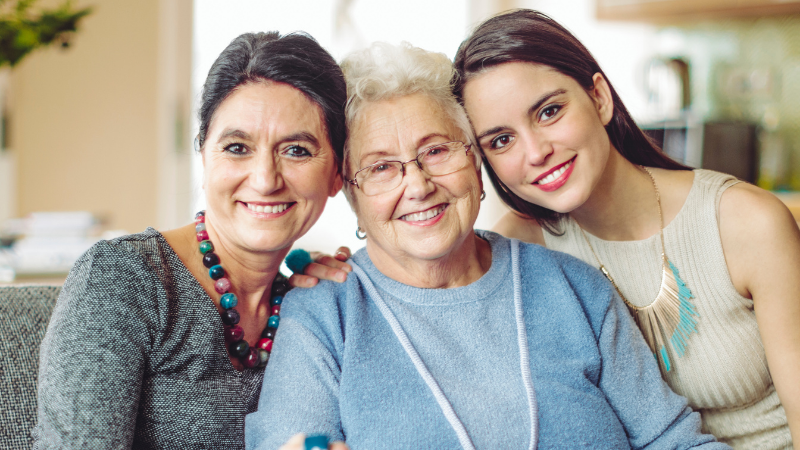 Three generations of women smiling on a sofa in a bright living room, arm in arm.