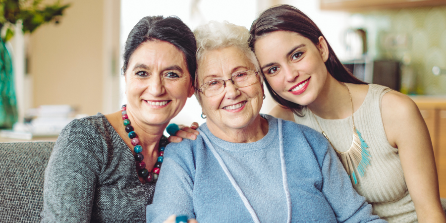 AccuQuote Blog 4 - AccuQuote Three generations of women smiling on a sofa in a bright living room, arm in arm.