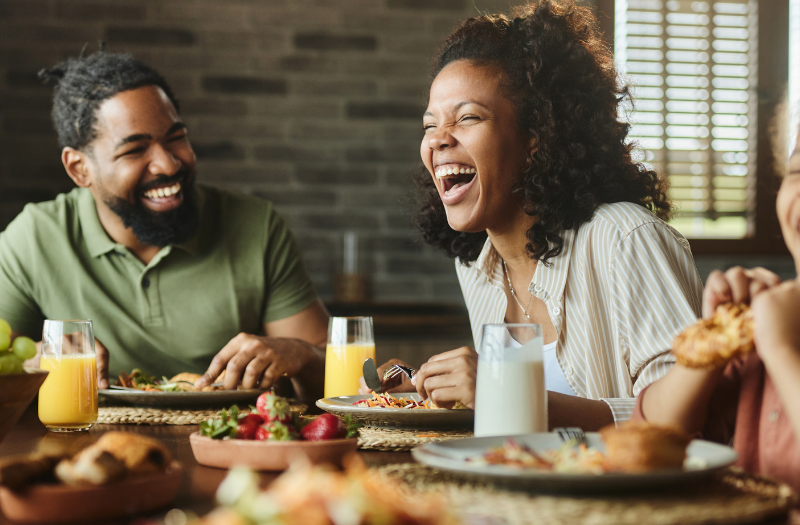 Three friends/family members laughing together at a dining table with plates, fruit bowl, and drinks on the table.