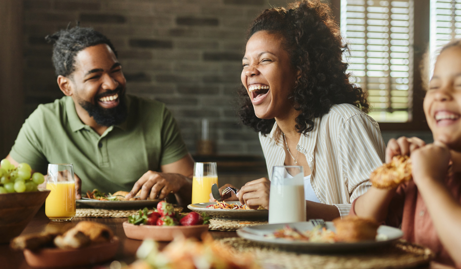 Three friends/family members laughing together at a dining table with plates, fruit bowl, and drinks on the table.