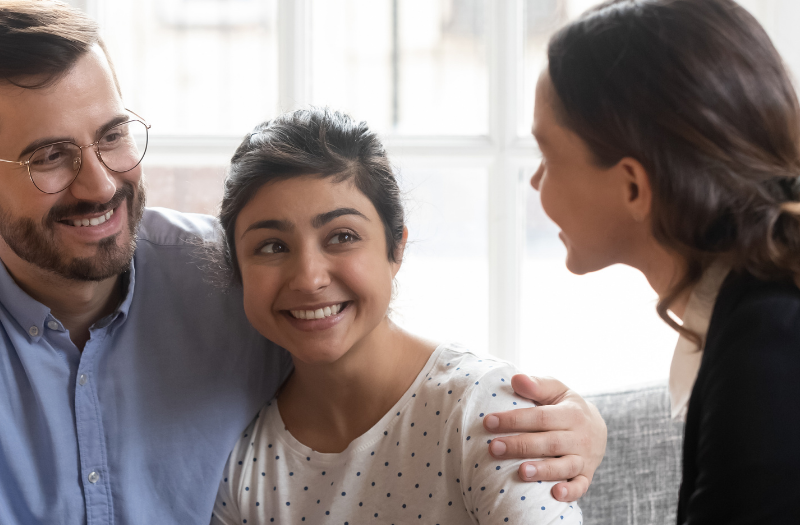 Three adults sitting on a couch, smiling and chatting in a bright living room.