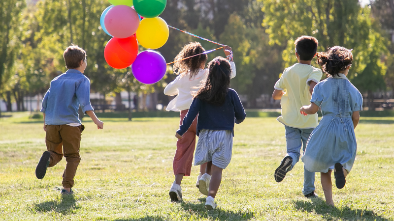 Five children in casual clothes run across a sunlit park, carrying a bundle of colorful balloons.