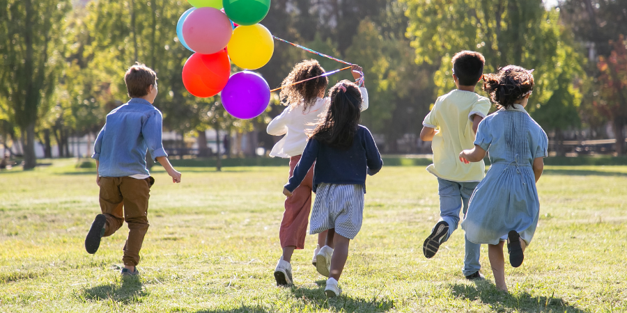 Five children in casual clothes run across a sunlit park, carrying a bundle of colorful balloons.
