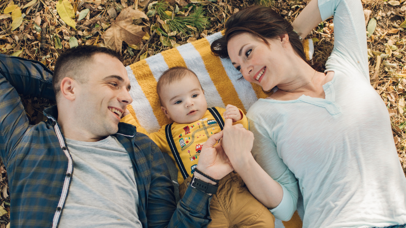 Family of three lying on a striped blanket outdoors, smiling at each other on autumn leaves.