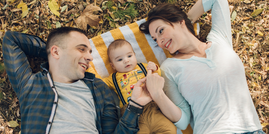 Family of three lying on a striped blanket outdoors, smiling at each other on autumn leaves.