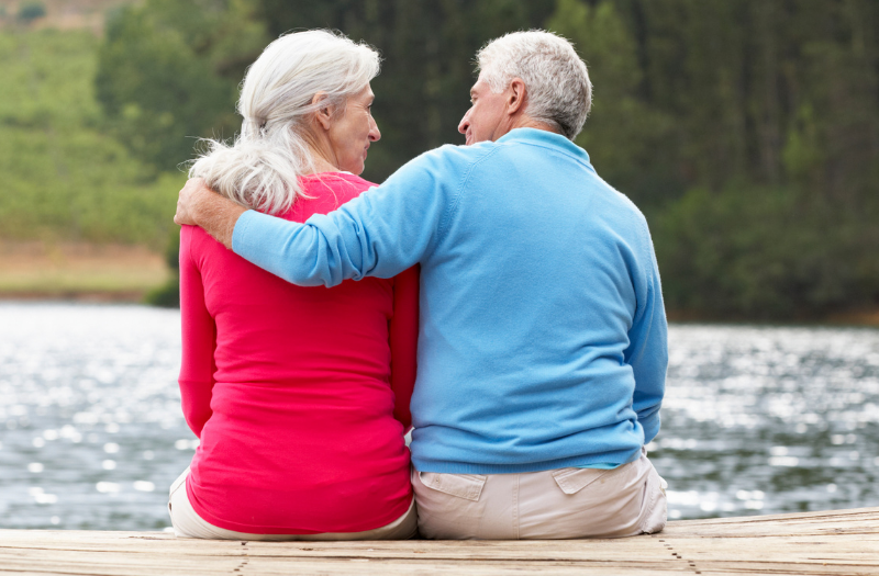 Older couple sits on a wooden dock by a lake, with arms around each other, enjoying a quiet moment.