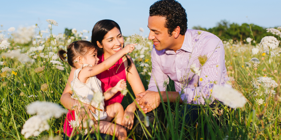 Family in a sunny wildflower field shares a moment of joy, daughter playing with parents nearby