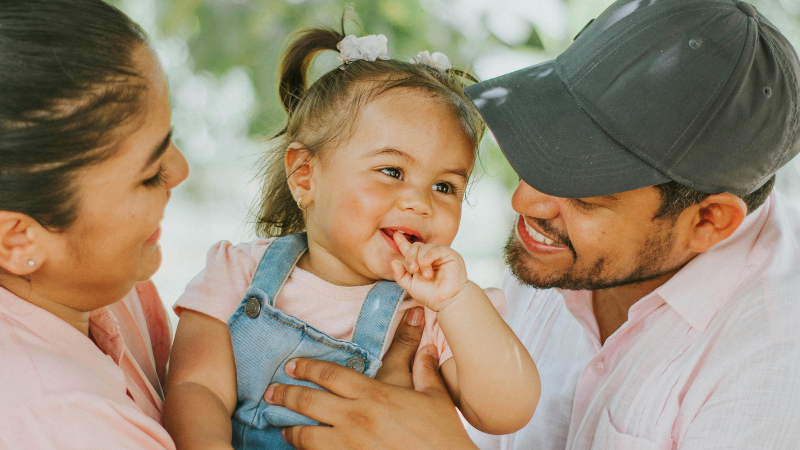 Happy family outdoors: mother and father smiling at their baby daughter with pigtails in a pink shirt and denim overalls.