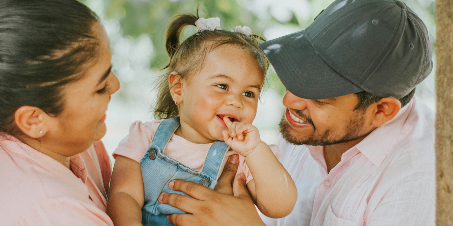Happy family outdoors: mother and father smiling at their baby daughter with pigtails in a pink shirt and denim overalls.