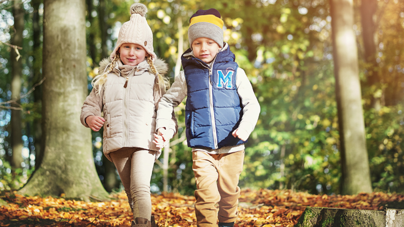 Two children walk hand in hand through a sunlit autumn forest carpeted with fallen leaves, wearing warm jackets and hats.