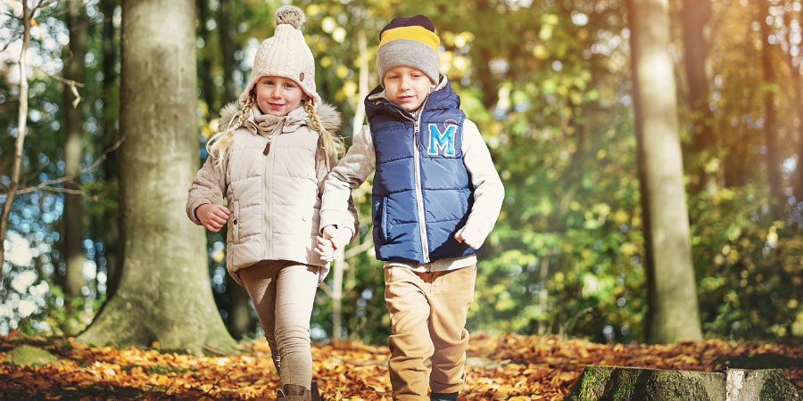 Two children walk hand in hand through a sunlit autumn forest carpeted with fallen leaves, wearing warm jackets and hats.
