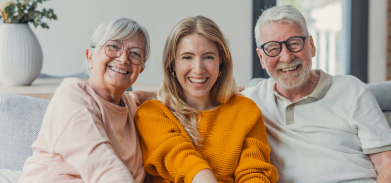 Three people smiling on a light gray sofa: an older woman in pink, a young woman in an orange sweater, and an older man with glasses nearby.