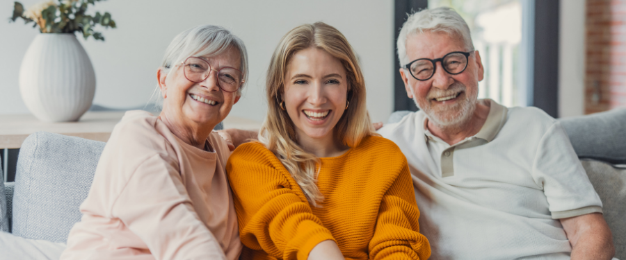 Three people smiling on a light gray sofa: an older woman in pink, a young woman in an orange sweater, and an older man with glasses nearby.
