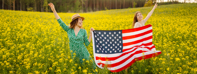 Two women stand in a sunny yellow flower field, waving an American flag.