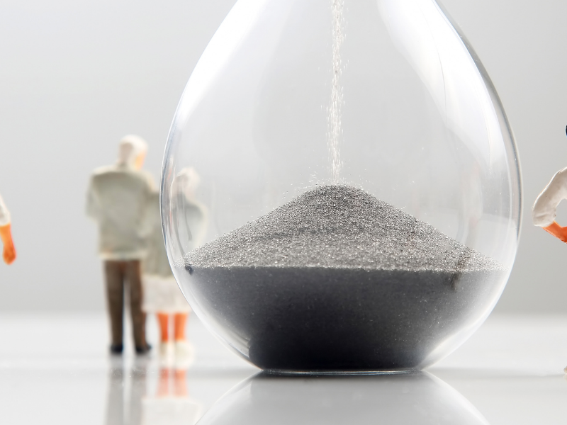 Miniature people gathered around a glass hourglass filled with sand, symbolizing time flowing amid a white surface.