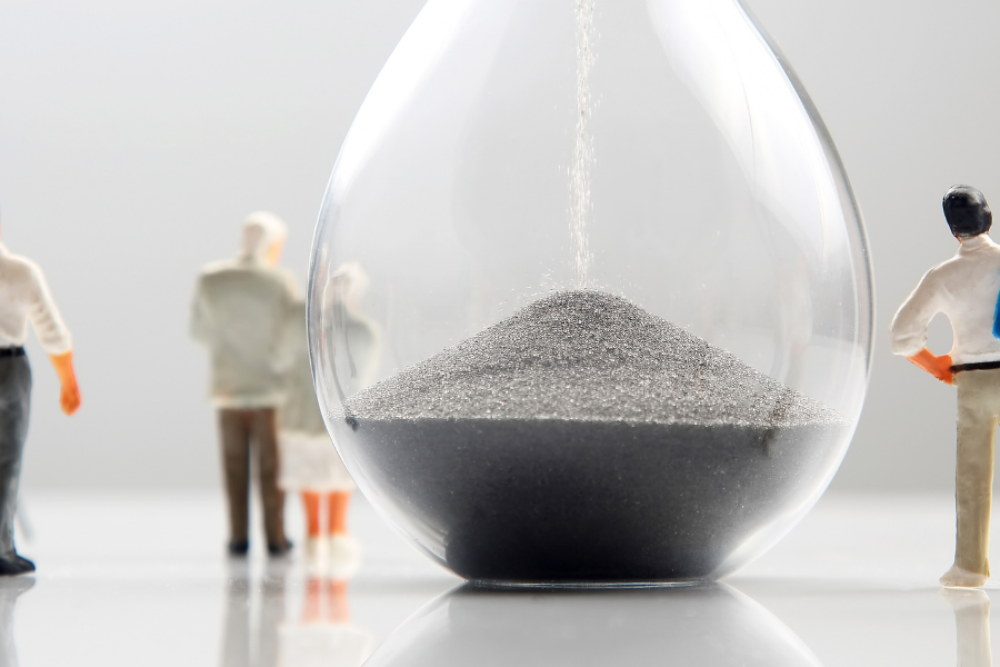 Miniature people gathered around a glass hourglass filled with sand, symbolizing time flowing amid a white surface.