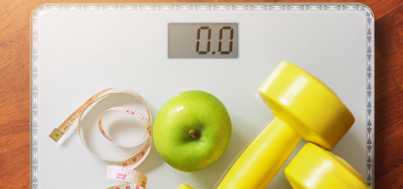 Scale shows 0.0 with a green apple, a measuring tape, and yellow dumbbells on a wooden floor, suggesting a fitness/weight goal scene.