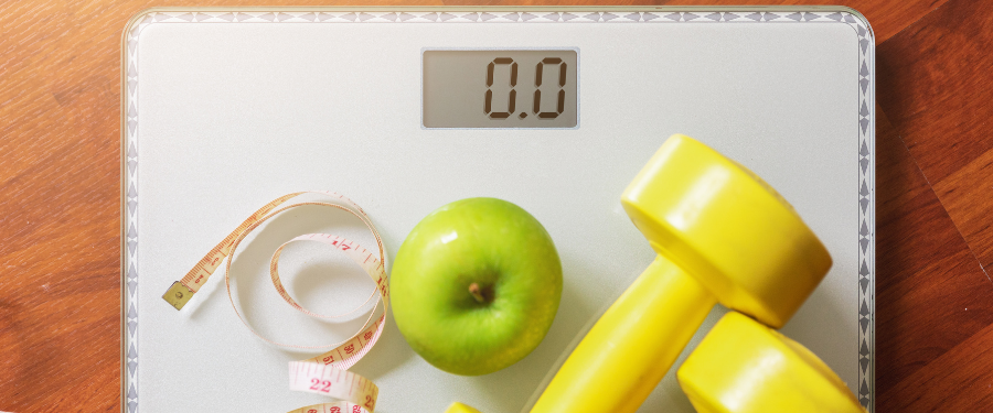 Scale shows 0.0 with a green apple, a measuring tape, and yellow dumbbells on a wooden floor, suggesting a fitness/weight goal scene.