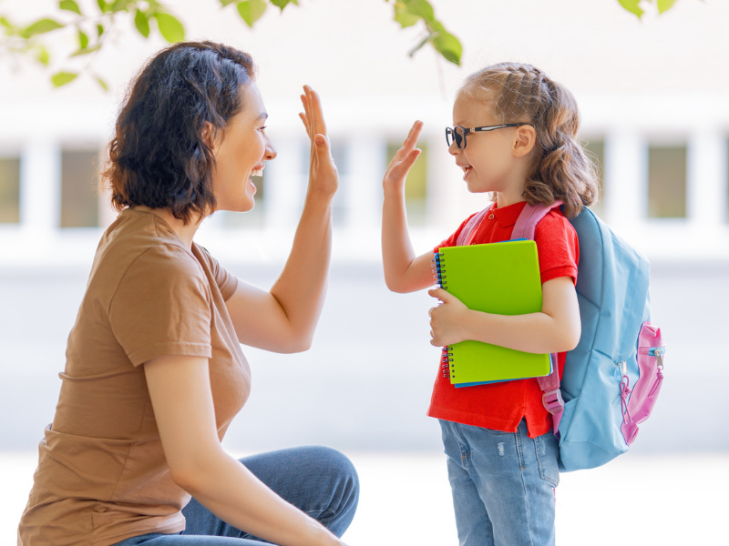 Teacher kneels to high-five a smiling student outdoors; student holds a bright green notebook and wears a blue backpack.