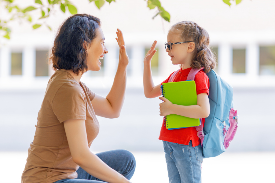 Teacher kneels to high-five a smiling student outdoors; student holds a bright green notebook and wears a blue backpack.