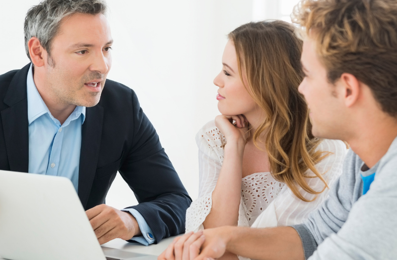 Business meeting: man in a suit explains something to a couple across an open laptop on a bright desk.