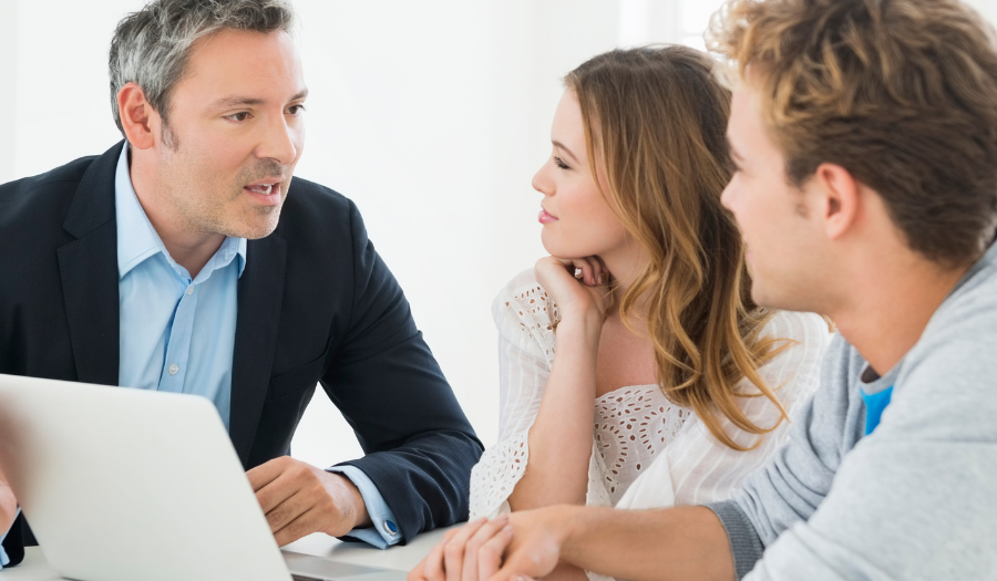 Business meeting: man in a suit explains something to a couple across an open laptop on a bright desk.