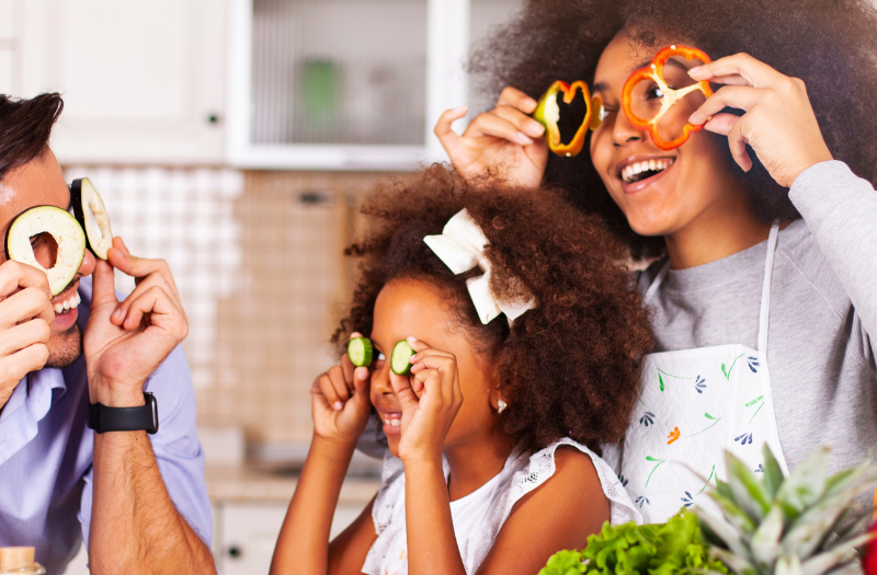 A family of three wearing vegetable slices as goggles and laughing in a bright kitchen with fresh produce nearby.