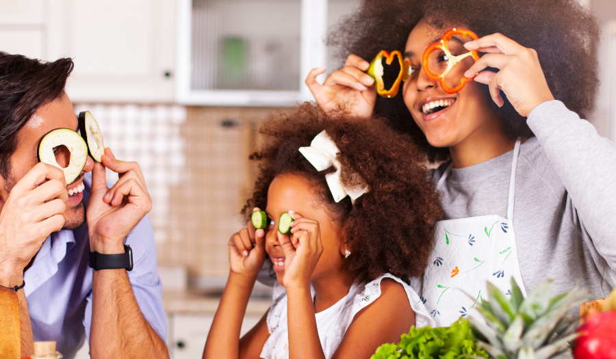 A family of three wearing vegetable slices as goggles and laughing in a bright kitchen with fresh produce nearby.