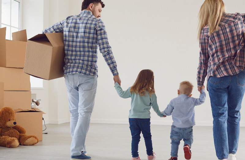 Family of four carrying moving boxes through a bright, empty room, holding hands with their children; a teddy bear sits on the floor near boxes.