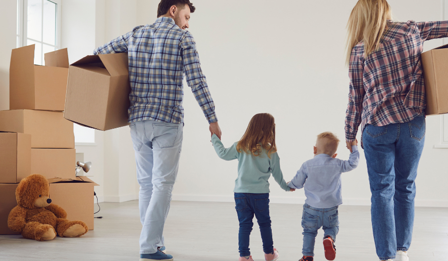Family of four carrying moving boxes through a bright, empty room, holding hands with their children; a teddy bear sits on the floor near boxes.