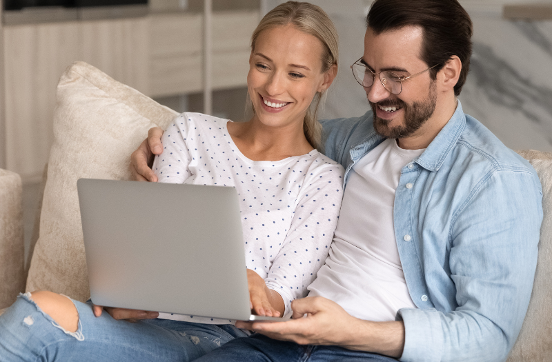 Couple relaxing on a beige sofa, looking at a laptop together and smiling at the screen in a home setting.