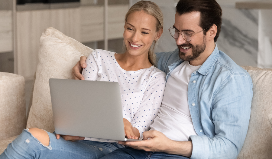 Couple relaxing on a beige sofa, looking at a laptop together and smiling at the screen in a home setting.