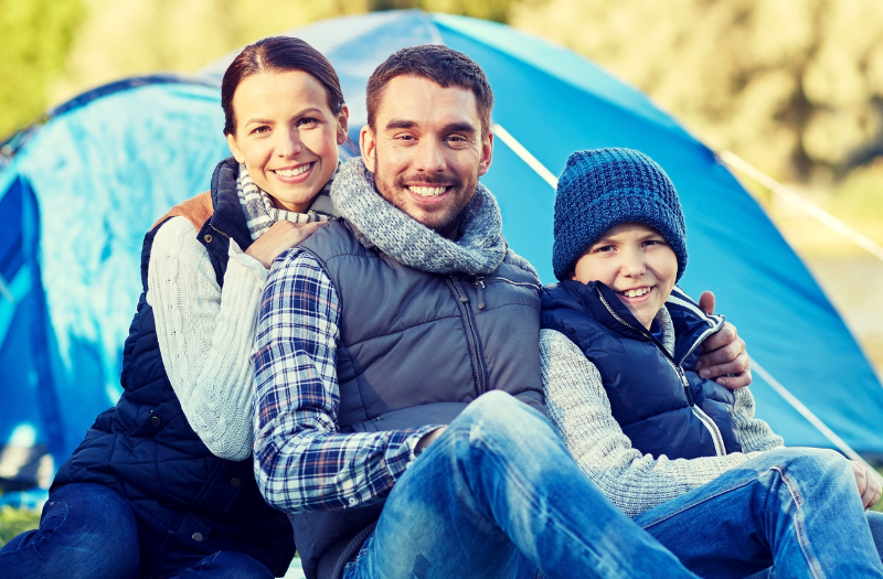 Family of three sits on the grass in front of a blue camping tent by a lake, smiling for the camera.