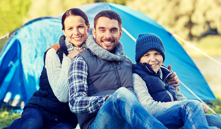 Family of three sits on the grass in front of a blue camping tent by a lake, smiling for the camera.