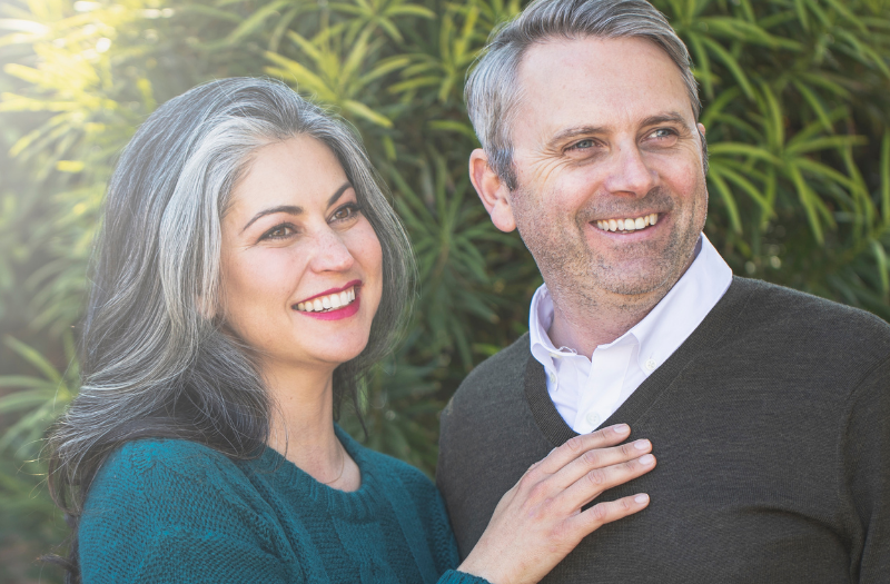 Smiling couple standing outdoors in greenery, bathed in sunlight on the left.