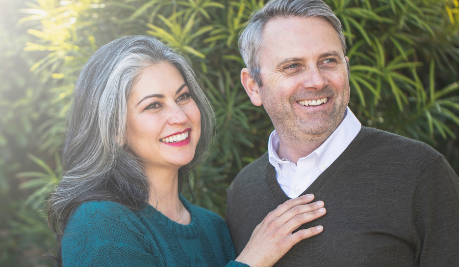 Smiling couple standing outdoors in greenery, bathed in sunlight on the left.
