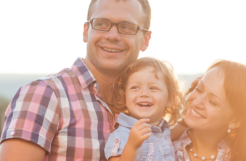 A smiling family of three hugs outdoors at sunset, mother and father with their toddler son or daughter.