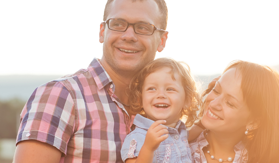 A smiling family of three hugs outdoors at sunset, mother and father with their toddler son or daughter.