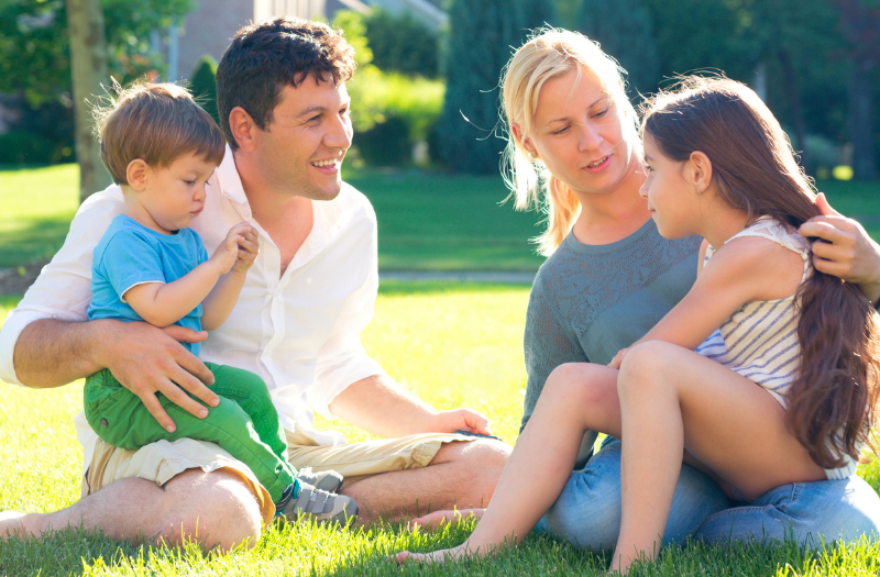 A family sits on a sunny lawn in a park, talking and smiling together with two young children on laps and nearby.
