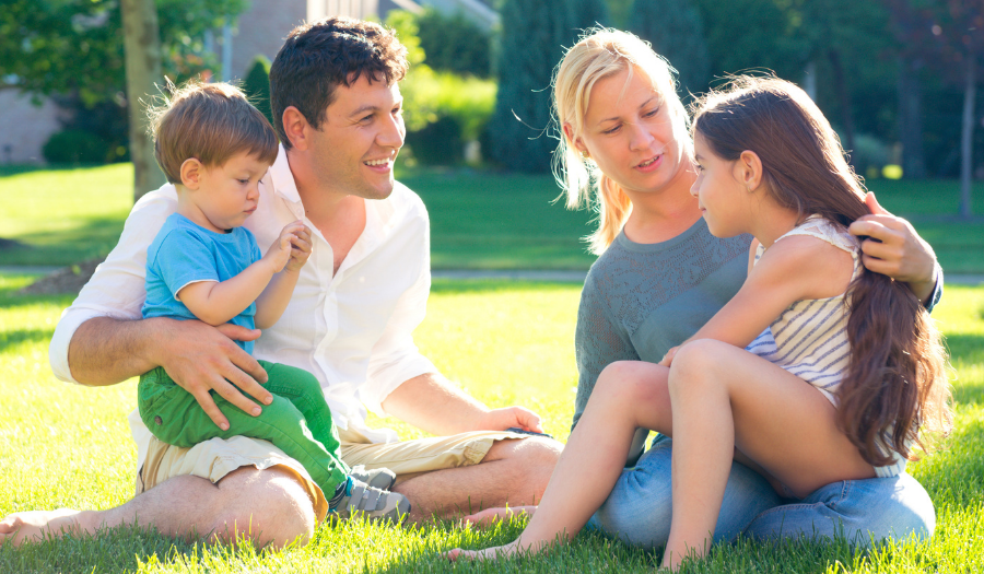 A family sits on a sunny lawn in a park, talking and smiling together with two young children on laps and nearby.