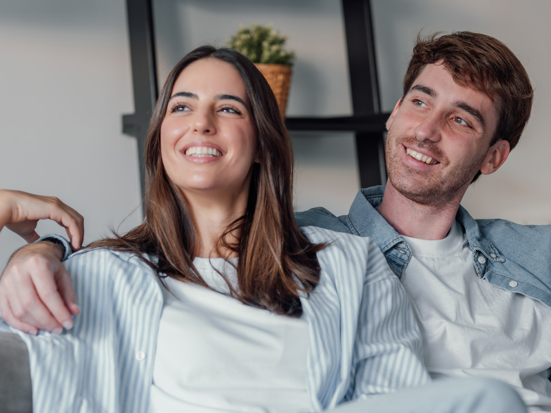 A happy couple relaxing on a sofa together, smiling and leaning on each other in a living room setting by the cushions on a gray couch.