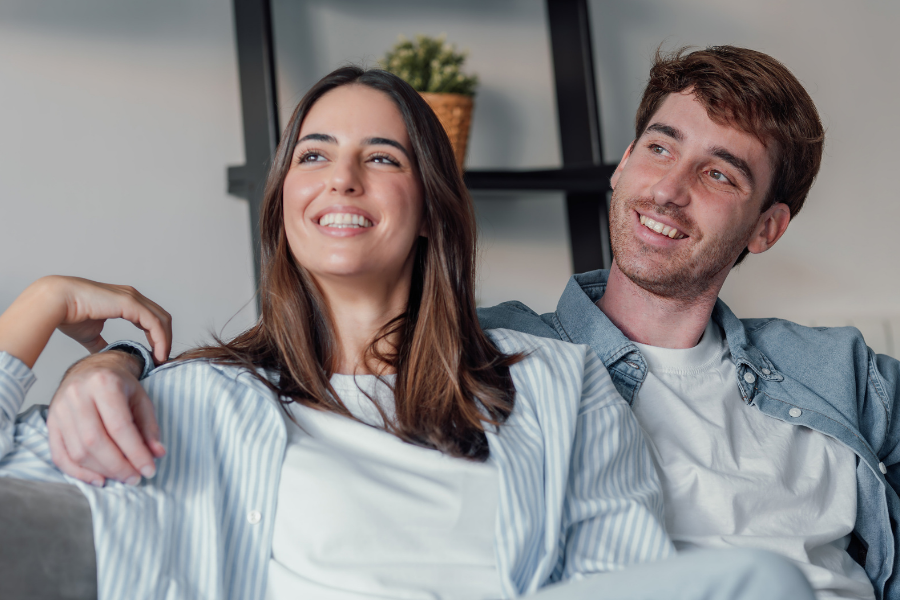 A happy couple relaxing on a sofa together, smiling and leaning on each other in a living room setting by the cushions on a gray couch.